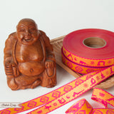 2 Webbandrollen mit dem Sanskritzeichen "Om" und eine mit Yogafiguren in pink und warmen gelb liegen neben einer Buddafigur aus Holz